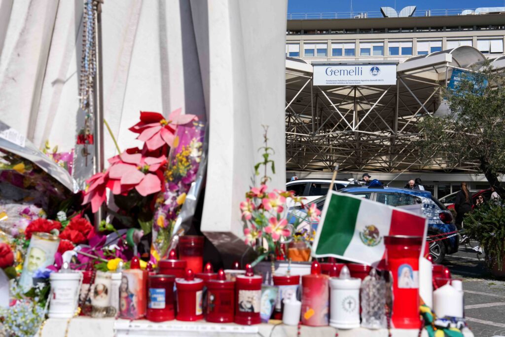 Velas votivas y flores en la base de una estatua de San Juan Pablo II fuera del hospital Gemelli de Roma el 6 de marzo de 2025, donde el Papa Francisco ha estado hospitalizado desde el 14 de febrero, recibiendo tratamiento para una neumonía doble. (CNS photo/Lola Gomez)