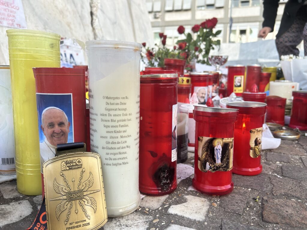 Una medalla de participante en el maratón de Roma se coloca entre las velas votivas y las flores depositadas en la base de la estatua de San Juan Pablo II, frente al hospital Gemelli de Roma, el 16 de marzo de 2025. El maratón de esa mañana comenzó con un momento de silencio para desear una pronta recuperación al Papa Francisco, que recibe tratamiento en el hospital desde el 14 de febrero. (Foto CNS/Pablo Esparza)