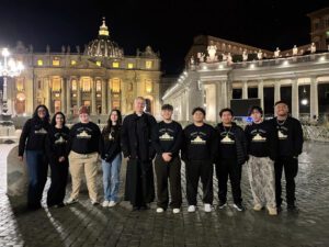 La clase de último año de nueve estudiantes de Holy Innocents School en Long Beach posa con el padre Robert McGowan en la Plaza de San Pedro durante la peregrinación del grupo a Italia. (Kiernan Fiore)
