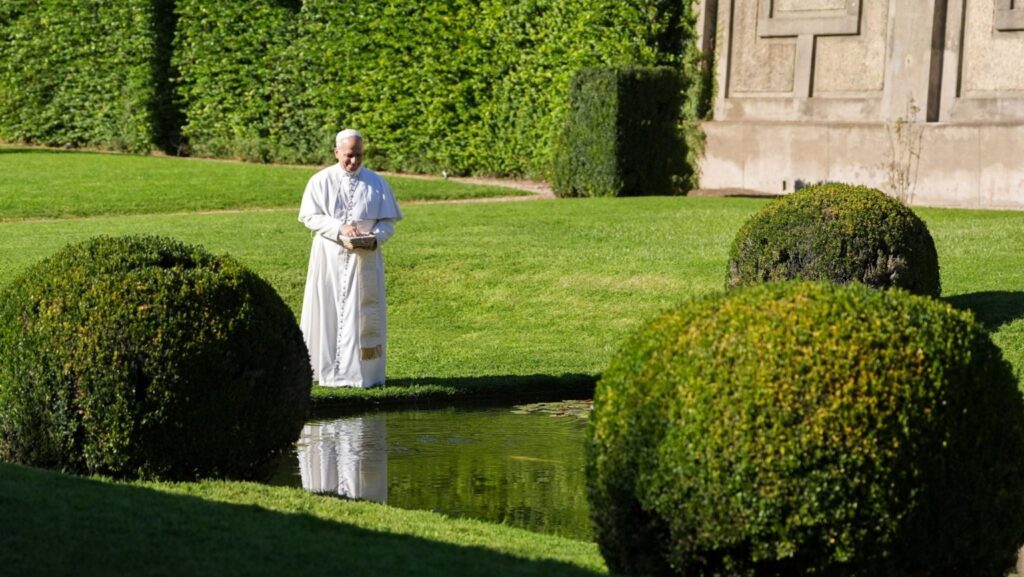 Man in white religious robes standing by a pond in a formal garden, holding a basket with a small item inside.