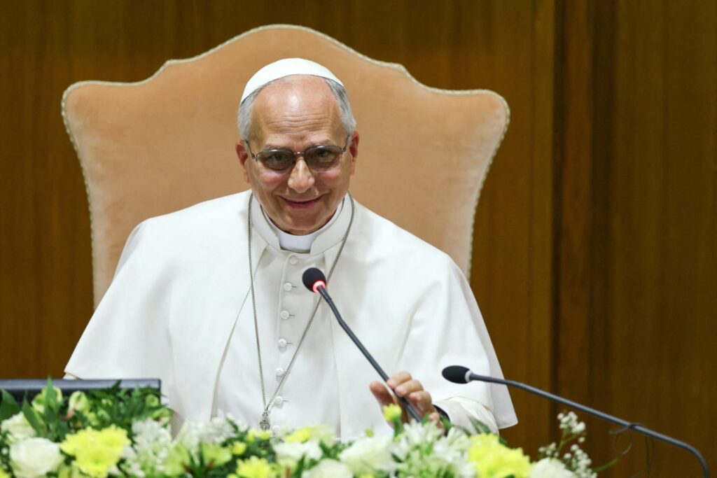Religious leader in white robes sits at a dais, smiling, speaking into a microphone, with a floral arrangement in front.
