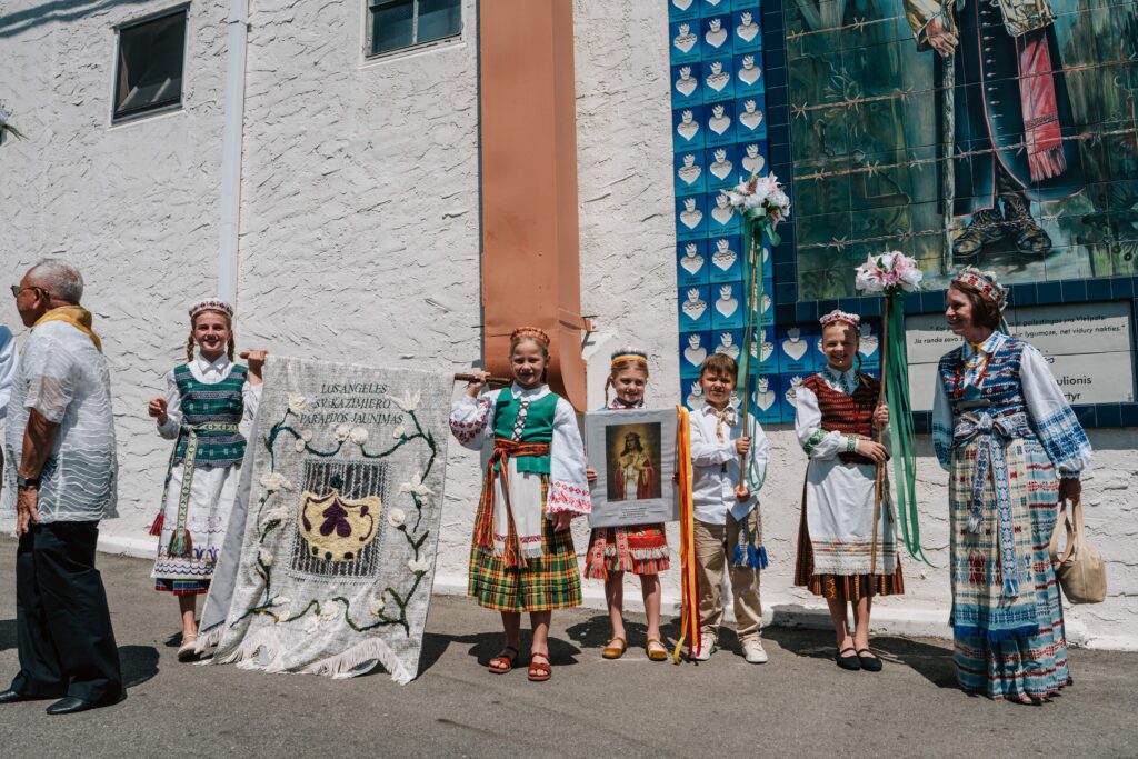 El padre Tomas Karanauskas, párroco de la Iglesia de San Casimiro en Los Feliz, posa en el centro con un saco rosa en honor a la tradicional sopa fría de remolacha lituana durante la celebración del aniversario Atlaidai de la parroquia, el 31 de mayo. A la izquierda aparece la secretaria parroquial, Vitalia Virbukiene, y a la derecha, la maestra de ceremonias del festival, Justina Brazdzionis. (Natalie Romano).