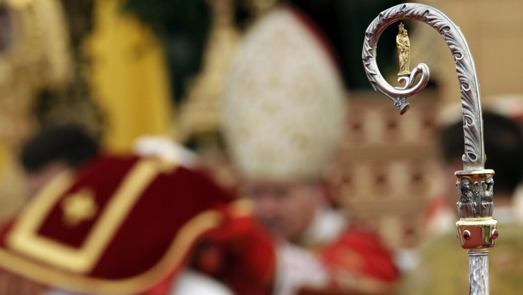 Close-up of an ornate ceremonial staff with a gold statue at the top, blurred red-and-gold uniforms in the background.