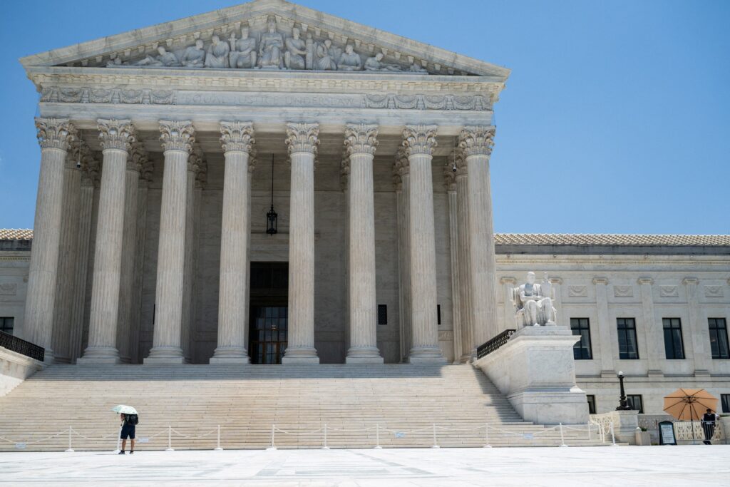 Marble neoclassical temple façade with tall Corinthian columns, wide stairs, and a seated statue on the right under a blue sky
