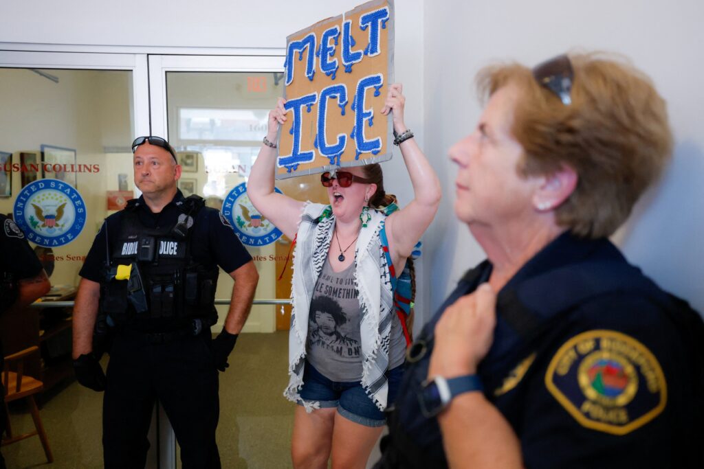 Una mujer sostiene un cartel frente a la oficina de la senadora republicana Susan Collins en Biddeford, Maine, el 13 de julio de 2026, mientras agentes de policía vigilan el lugar y varias personas se congregan para protestar después de que un agente del Servicio de Inmigración y Control de Aduanas (ICE) de Estados Unidos matara a tiros a un automovilista. (Foto de OSV News/CJ Gunther, Reuters)