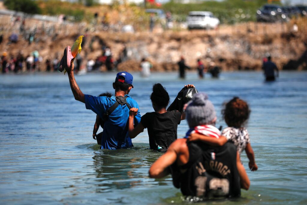 Group of people wearing life jackets wading through water toward a shore, with a rocky embankment and onlookers in the background.