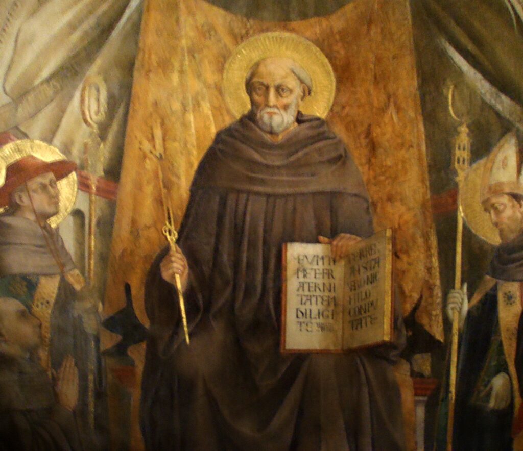 Statue of a bearded saint with a golden halo, dressed in brown robes, standing among other figures in a wooden display backdrop.