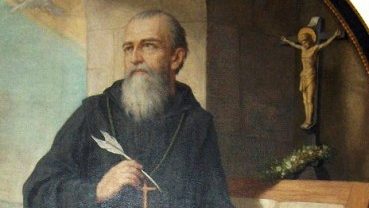 Portrait of a bearded monk or friar in dark robes, holding a quill near a cross on a desk with a crucifix in the background.