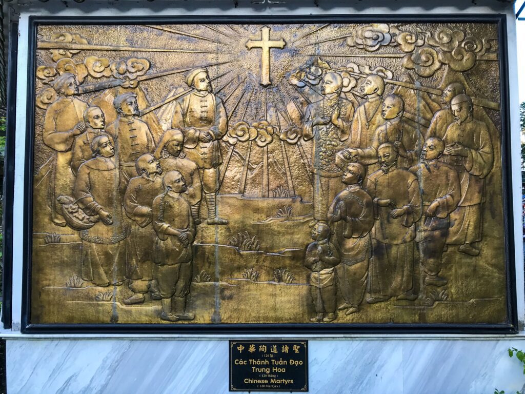 Bronze relief panel depicting a crowd of Chinese martyrs surrounding a central cross, with radiant lines and cloud motifs above.