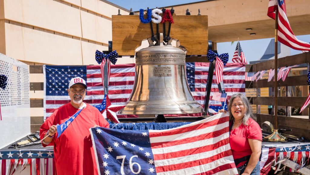 Two people pose beside a large silver bell on a patriotic float decorated with American flags and red‑white‑blue bunting.
