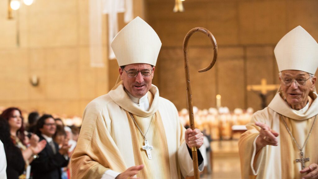 Two Catholic bishops in cream robes and white mitres walk in a church procession, one holding a wooden crozier as the crowd applauds in the background.