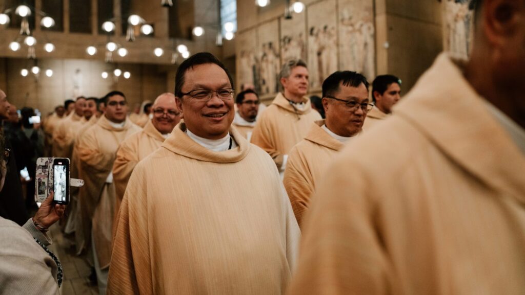 Monks in beige robes smile as they walk through a church, with a spectator recording them on a smartphone.