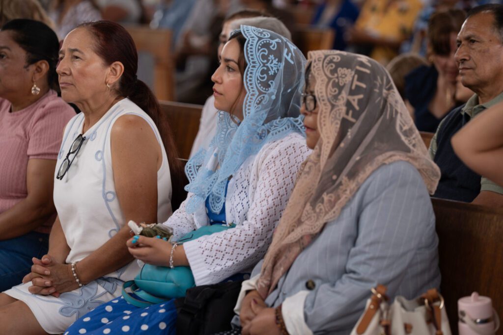 Three women sit in a row on wooden pews at a formal event: a woman in a white sleeveless dress with glasses hanging from her chest, a woman in a light blue lace headscarf, and a woman in a beige lace hijab.
