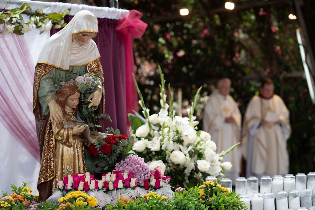 Statue of Mary holding baby Jesus at an outdoor altar, surrounded by white flowers and candles, with two clergymen in robes in the background.