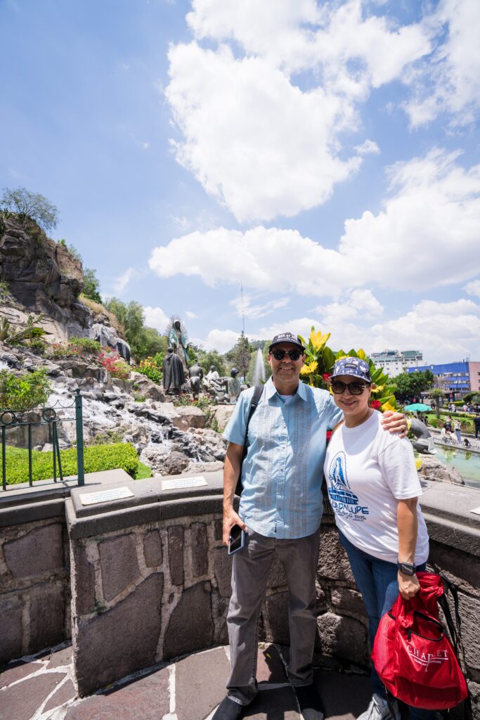 Smiling man and woman pose together outdoors near a rocky garden with statues under a blue sky.