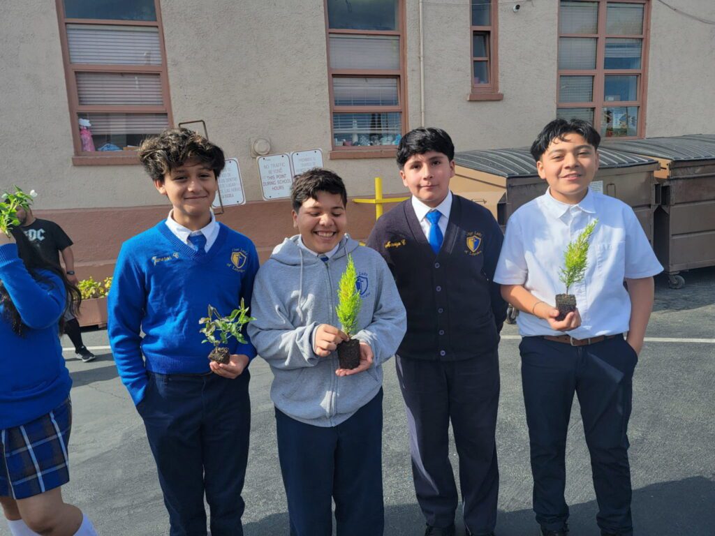 Four students in school uniforms hold small potted plants outdoors, smiling for a photo visit to a garden project.