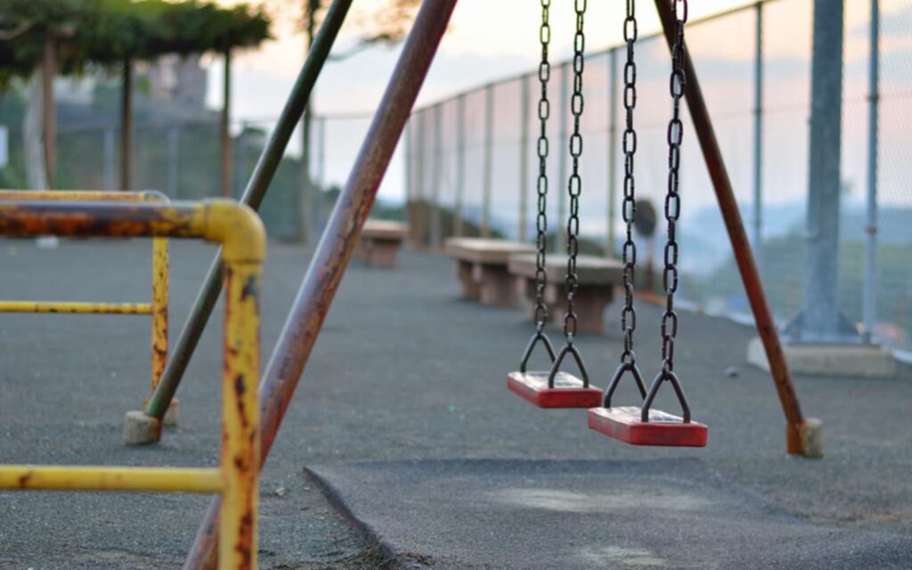 Three red swing seats on chains in a deserted playground, with rusted yellow support bars and a chain-link fence in the background.