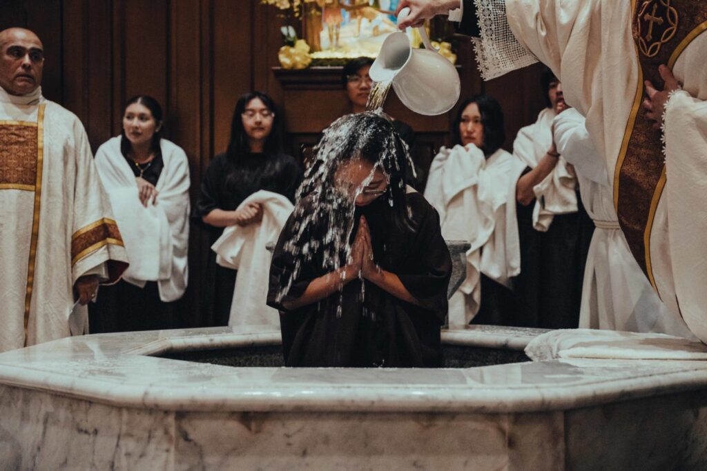 Person in dark robes receives water blessing as a priest pours water over their head while others in white robes watch with towels nearby at a baptism ceremony in a marble font area.