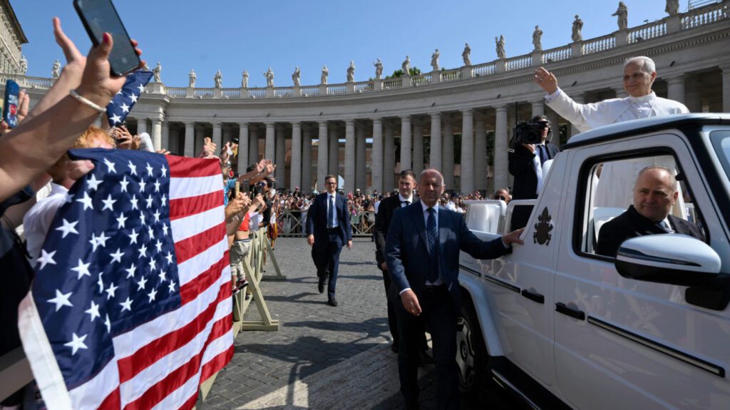 Religious leader in white robes waves from a white SUV as crowds cheer, with American flags held by supporters along a barrier in a sunny square.