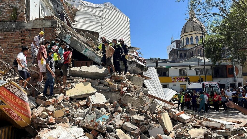 Rescue workers and bystanders on a collapsed building site, with rubble and debris on a street and a church in the background.