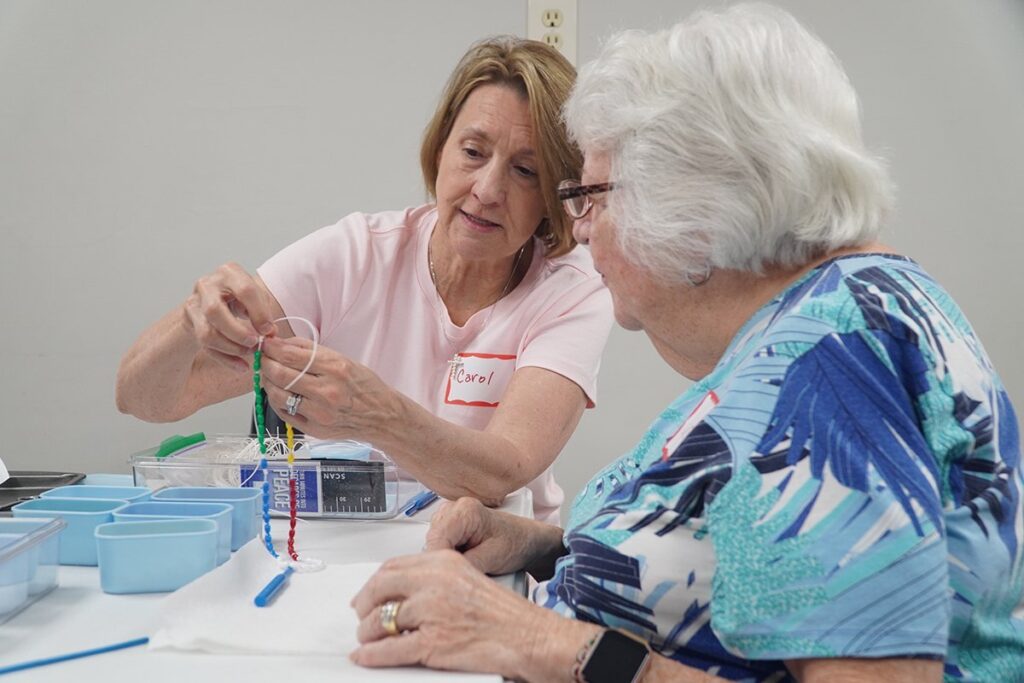Two senior women at a craft table stringing colorful beads; one has a name tag reading Carol.