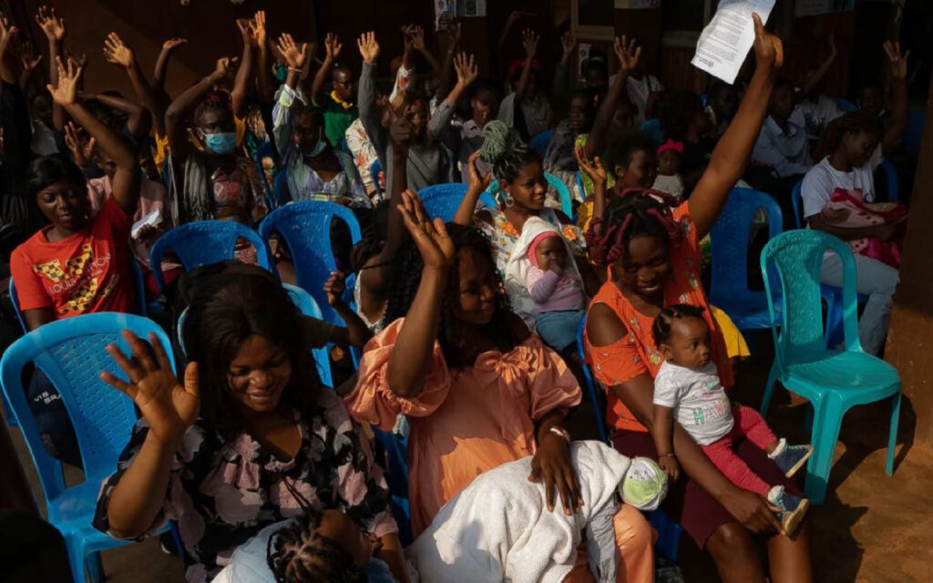 Crowded indoor gathering with many people raising hands; foreground woman holds a baby on her lap.