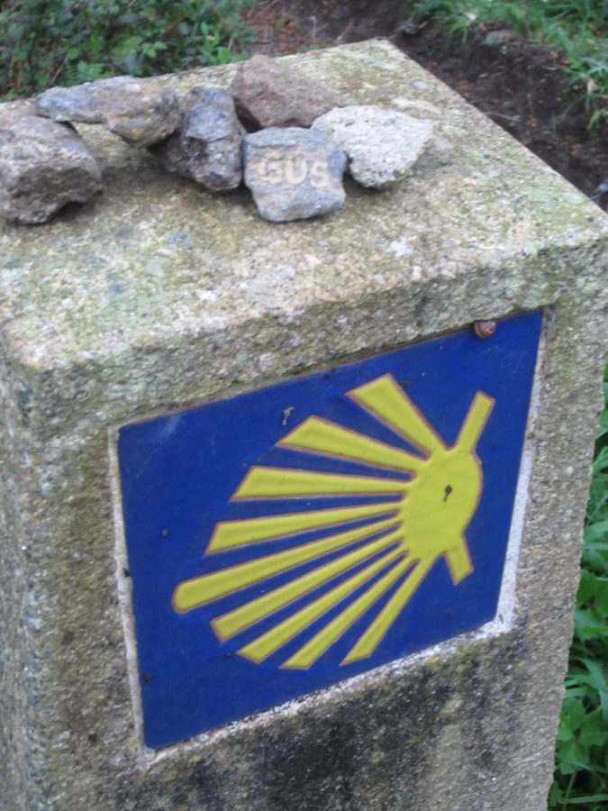 Stone milestone with a blue plaque showing a yellow scallop shell, a Camino de Santiago marker on a mossy concrete post.