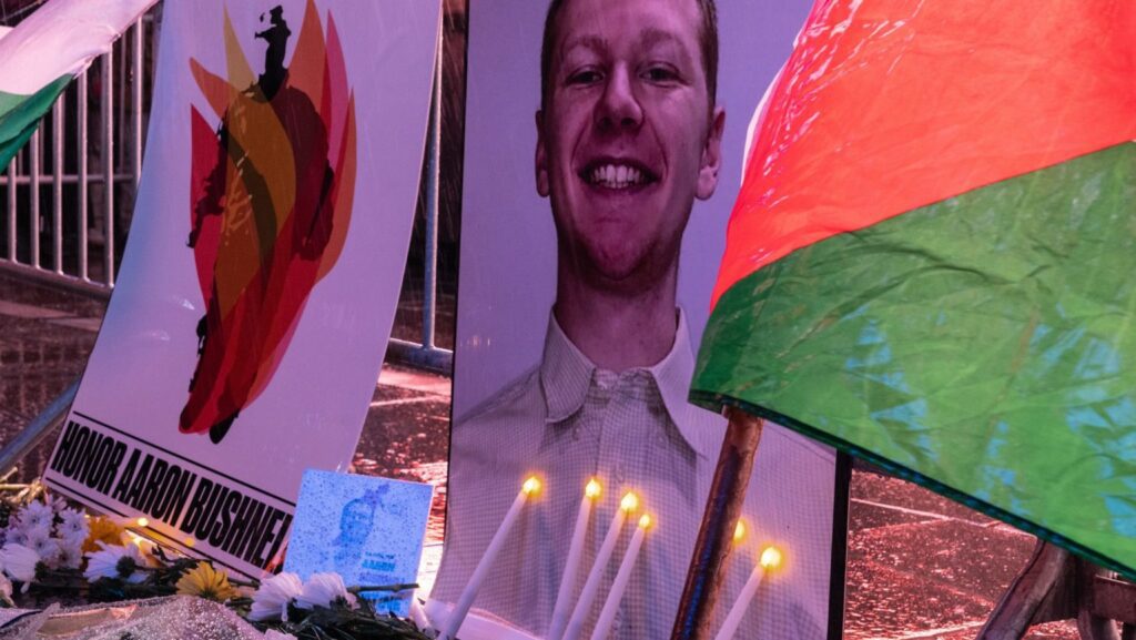 Memorial display with a large portrait of a smiling man, candles and flowers on a street, and a banner reading 'HONOR AARON...' in the background; colorful flags nearby.