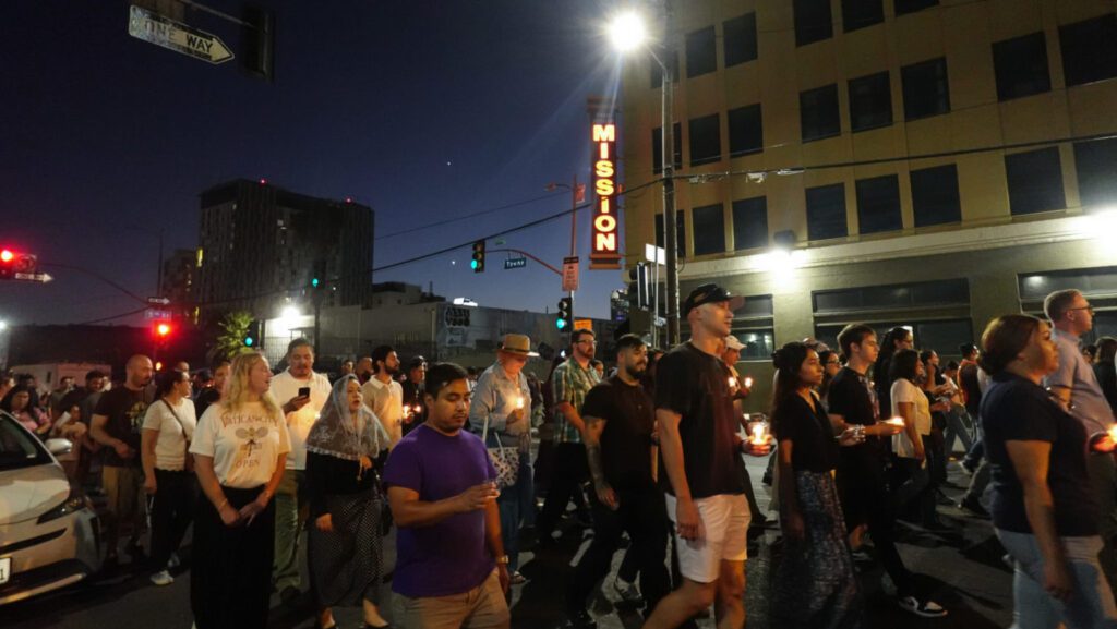 Diverse group of people walking down a city street at night, holding lit candles in a peaceful vigil under streetlights and a Mission sign on a building behind them.
